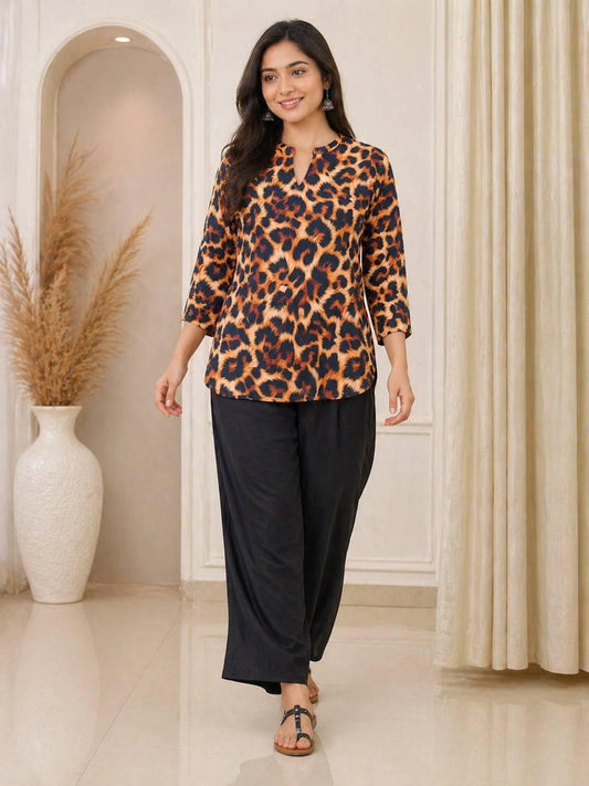 A woman modeling a leopard print top with three-quarter sleeves and black wide-leg pants, standing in a decorated indoor setting with beige walls, a large white vase with dried grass, and cream curtains.